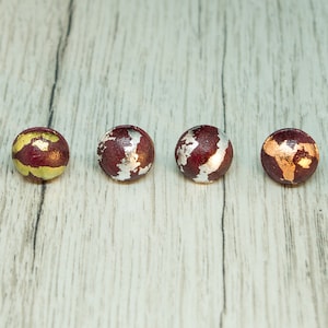 May include: Six round wooden earrings with a burgundy base and gold, silver, or copper accents. The earrings are arranged in a row on a light wood surface.
