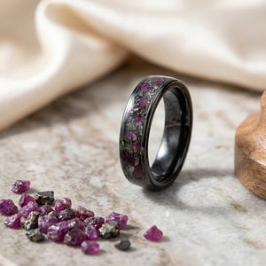 May include: A black ring featuring an inlay of crushed deep pink ruby fragments and metallic flecks. The band has a smooth, polished finish, and the center channel is filled with a textured, multi-colored stone and metal composite material.
