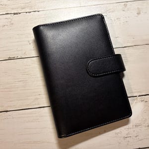 May include: A black leather journal with a snap closure. The journal is rectangular and has a smooth, matte finish. The cover is closed with a strap that fastens with a snap. The journal is on a light wood surface.