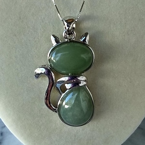 May include: A silver-toned cat pendant necklace with a green stone body and head. The cat pendant has a rounded shape with a tail and ears. The necklace is displayed on a neutral background.