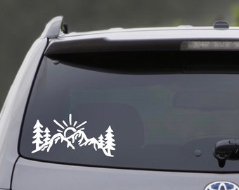 May include: White vinyl decal featuring a mountain range, sun, and trees. The design is a silhouette, suitable for application on a car window or other smooth surfaces. The decal is a simple, yet elegant design.