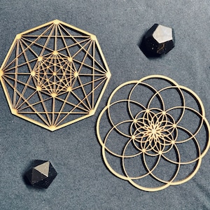 以下が含まれることがあります： 暗い青色の表面に、2つの木製の幾何学模様と2つの黒い宝石が配置されています。1つのデザインは、複雑な内部パターンを持つ多面的な形状です。もう1つは、重なり合う円の円形デザインです。