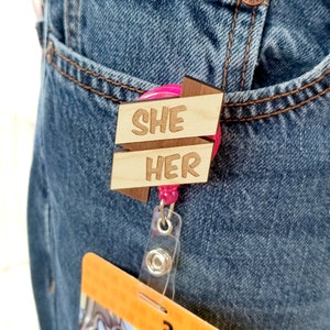 May include: A wooden badge holder with a pink retractable reel. The badge holder has two wooden arrows with the words "SHE" and "HER" written on them.