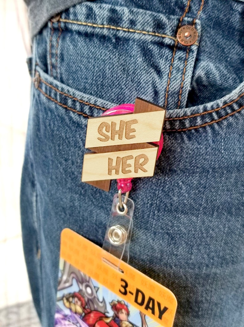May include: A wooden badge holder with a pink retractable reel. The badge holder has two wooden arrows with the words "SHE" and "HER" written on them.
