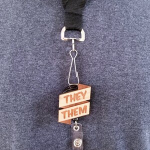May include: A black lanyard with a wooden badge holder that says "They Them" and a retractable badge holder with a 3-day pass.