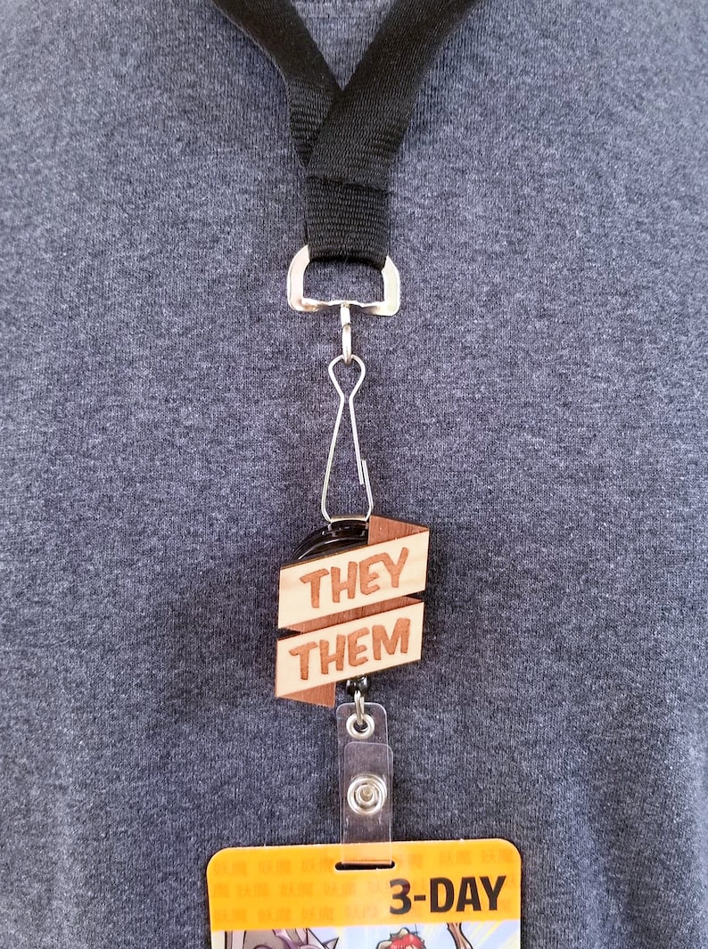 May include: A black lanyard with a wooden badge holder that says "They Them" and a retractable badge holder with a 3-day pass.