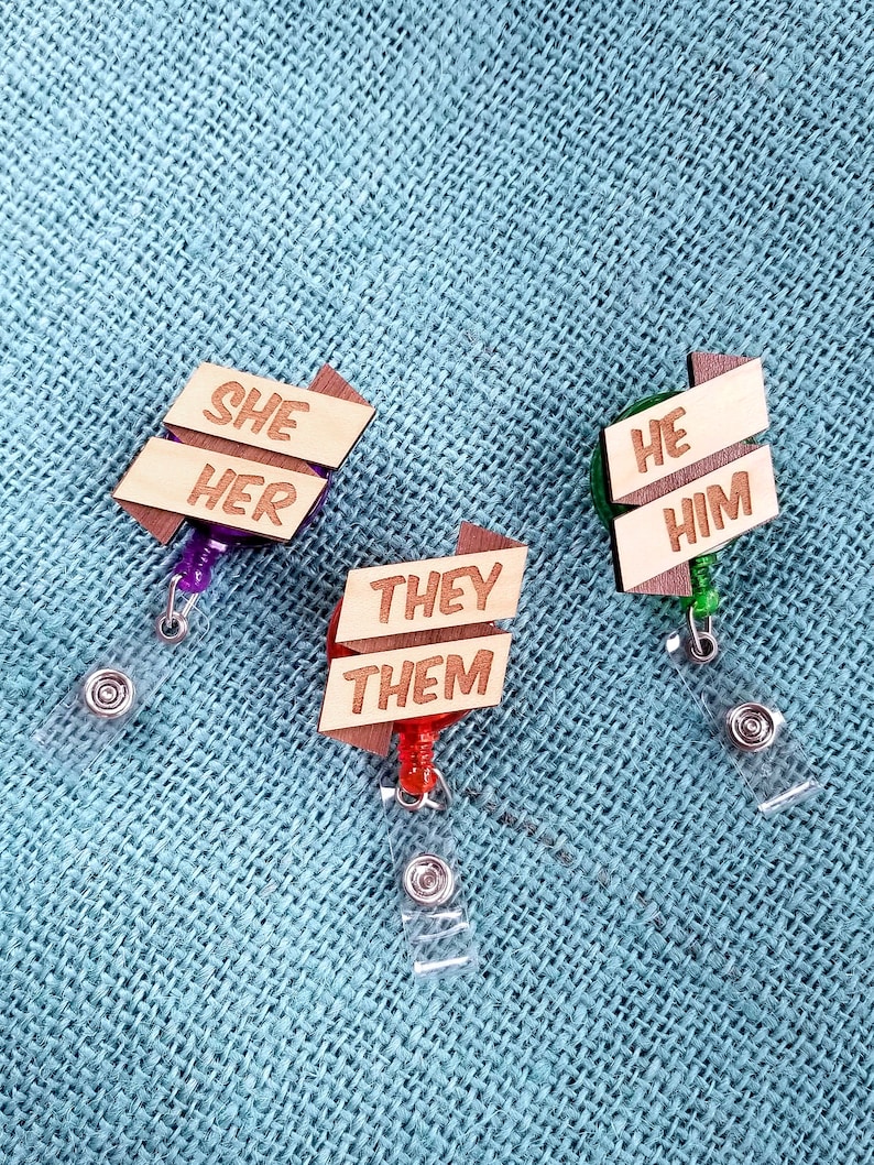 May include: Three wooden badge holders with retractable clips. The holders are shaped like arrows and have the words "She Her", "He Him", and "They Them" written on them.