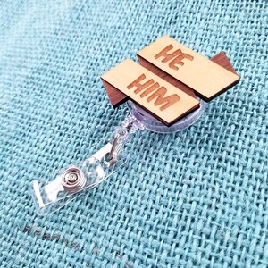 May include: A clear plastic retractable badge holder with a wooden tag attached. The tag is made of two pieces of wood, one with the word "HE" and the other with the word "HIM".