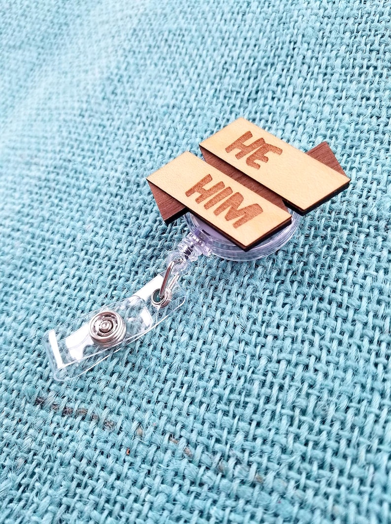 May include: A clear plastic retractable badge holder with a wooden tag attached. The tag is made of two pieces of wood, one with the word "HE" and the other with the word "HIM".