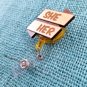 May include: A wooden badge holder with a yellow retractable reel. The badge holder has two wooden signs attached to it. The top sign says "SHE" and the bottom sign says "HER".