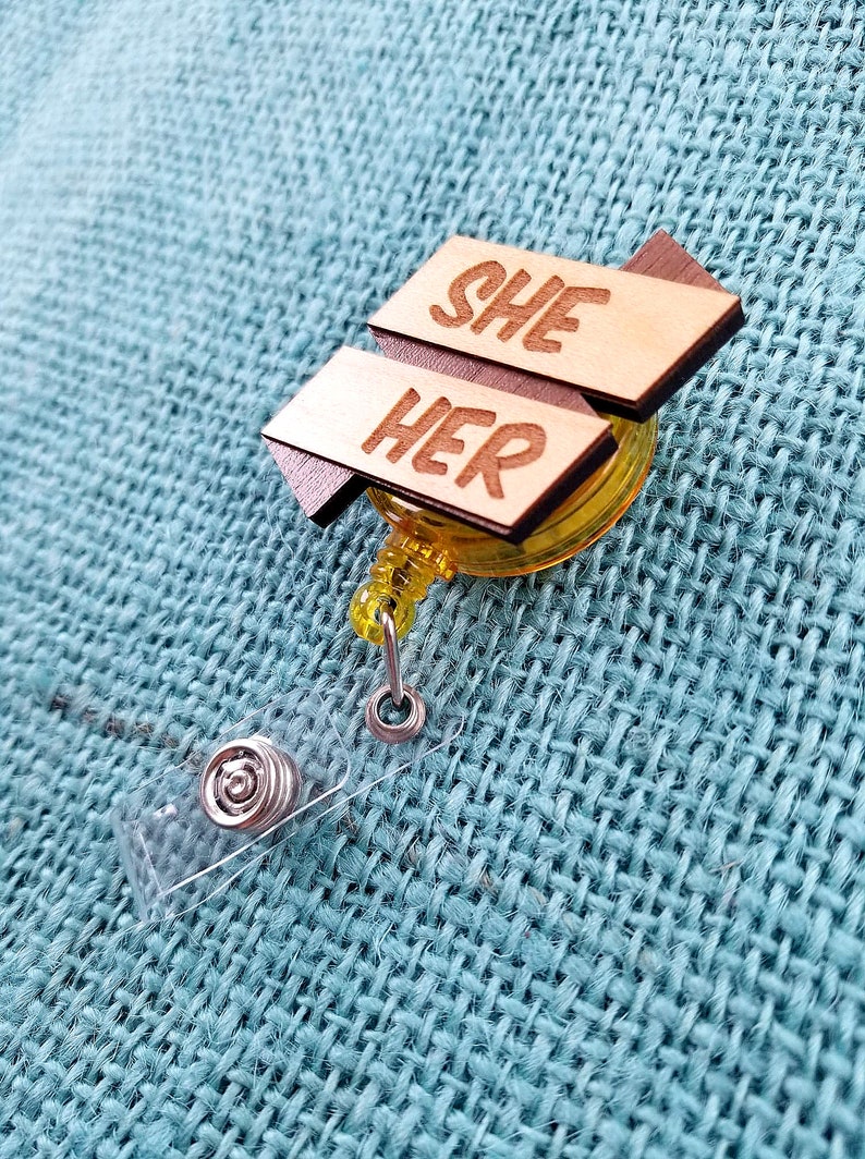 May include: A wooden badge holder with a yellow retractable reel. The badge holder has two wooden signs attached to it. The top sign says "SHE" and the bottom sign says "HER".