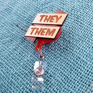 May include: A wooden badge holder with a red retractable reel. The wooden sign reads "They Them" in all caps.