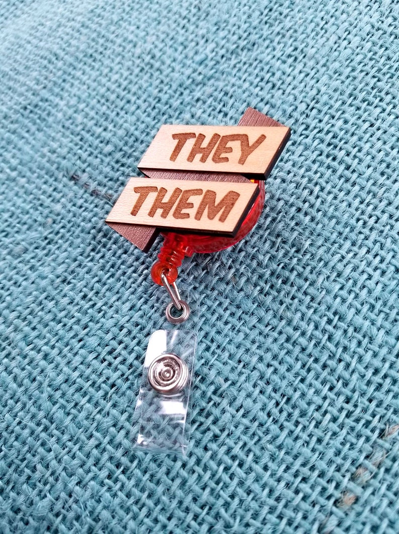 May include: A wooden badge holder with a red retractable reel. The wooden sign reads "They Them" in all caps.