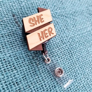 May include: Wooden badge holder with a retractable cord. The badge features two wooden signs with the words "SHE" and "HER" engraved on them.