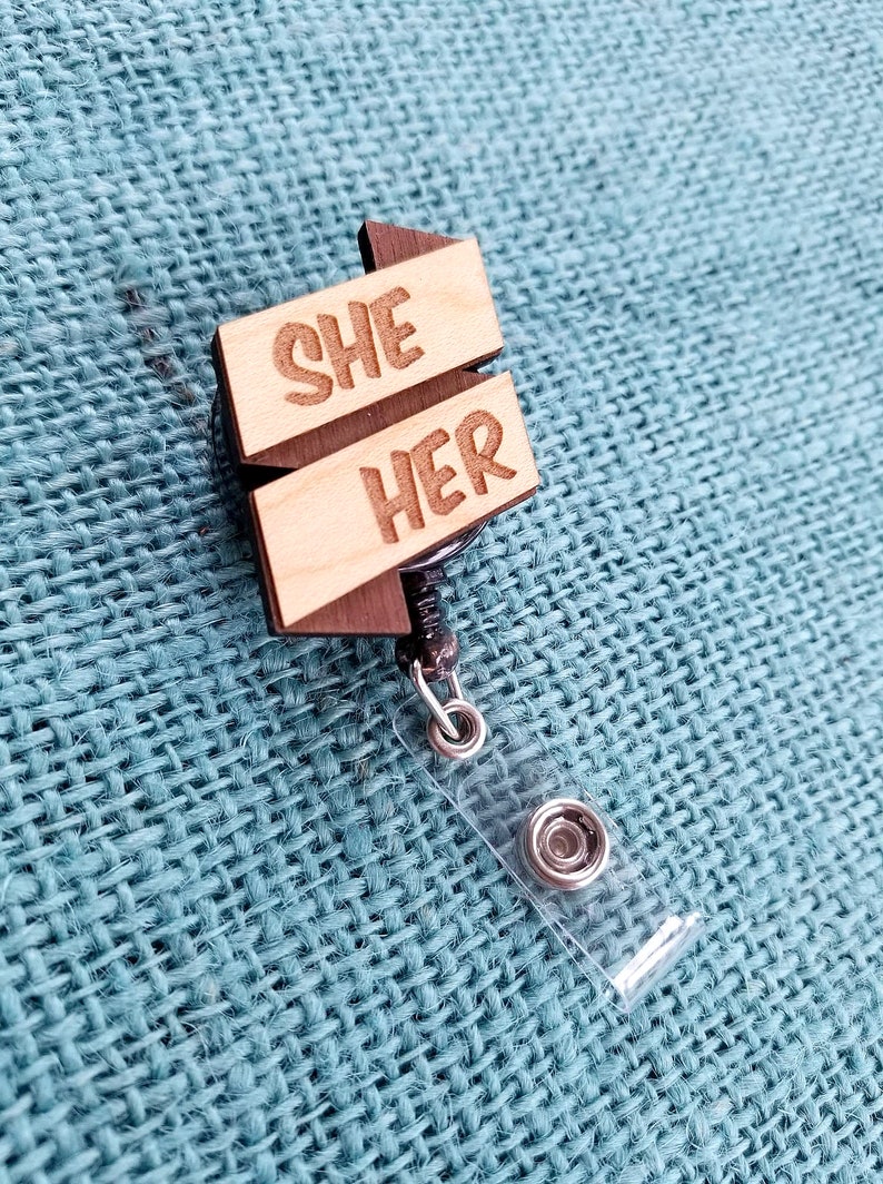 May include: Wooden badge holder with a retractable cord. The badge features two wooden signs with the words "SHE" and "HER" engraved on them.