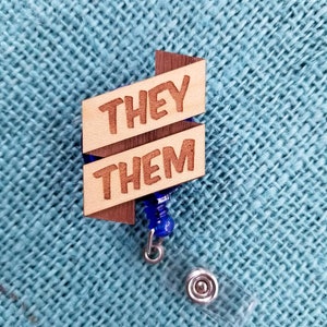 May include: Wooden badge holder with a blue retractable reel. The badge holder has two wooden pieces with the words "THEY" and "THEM" engraved on them.