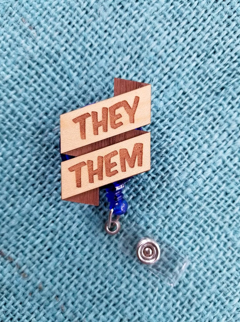 May include: Wooden badge holder with a blue retractable reel. The badge holder has two wooden pieces with the words "THEY" and "THEM" engraved on them.