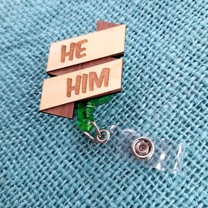 May include: Wooden badge holder with a green retractable cord. The badge has the words "HE HIM" written on it.