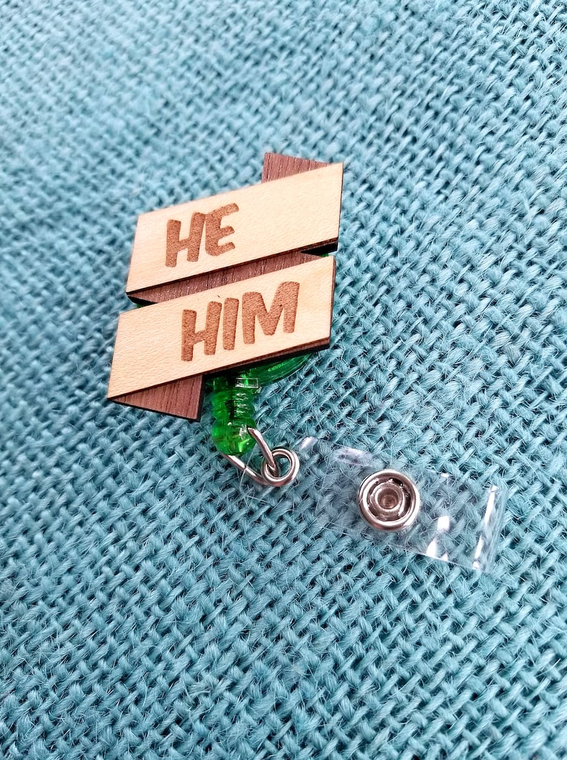 May include: Wooden badge holder with a green retractable cord. The badge has the words "HE HIM" written on it.
