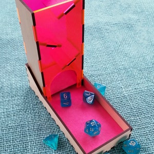 May include: A wooden dice tower with a pink acrylic insert and a pink tray. The tower has a slot for dice to fall through. There are six blue dice with gold numbers on a pink tray. The dice are a d4, d6, d8, d10, d12, and d20.