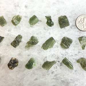 May include: A collection of 17 green Moldavite crystals of various sizes and shapes. The crystals are arranged on a white surface. A US dime coin is included for scale.