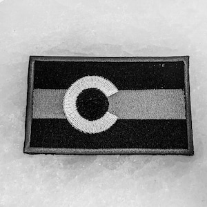 Puede incluir: Un parche bordado en blanco y negro con la bandera del estado de Colorado. La bandera tiene un círculo blanco con una 'C' negra en su interior, y tres franjas horizontales: una franja gris claro, una franja gris oscuro y una franja negra.