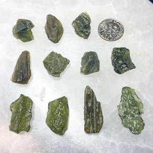 May include: A collection of 12 green aventurine crystals of varying sizes and shapes. The crystals are arranged on a white surface. A 10 pence coin is included for scale.