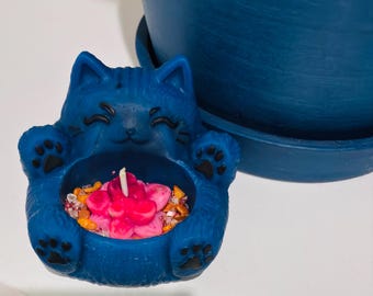 May include: A blue cat-shaped candle holder with black painted details on the eyes and paws. The centre contains a pink flower-shaped candle surrounded by small, multi-coloured decorative stones and a single white wick in the middle.