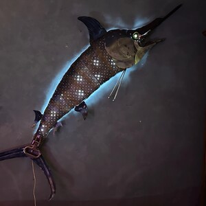 May include: A metal sculpture of a marlin fish with a blue glow and a starry pattern. The sculpture is mounted on a wall.