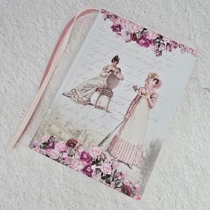 May include: A pink and white journal with a ribbon bookmark. The cover features a vintage illustration of two women in dresses with a floral border. The text on the cover reads "What I have learned in the journey of life..."