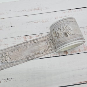 May include: A roll of decorative trim with a distressed, off-white finish. The trim features embossed floral and leaf designs, adding a vintage or antique aesthetic. The trim is likely for home decor or craft projects.