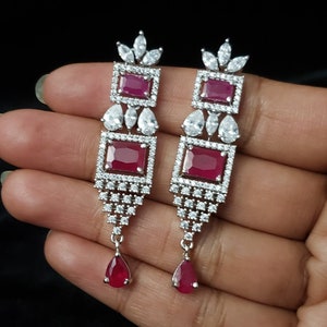 May include: A pair of dangle earrings with a teardrop-shaped red stone at the bottom. The earrings are made of silver and have a cluster of clear stones in the shape of a flower at the top. The earrings have three rectangular red stones in the middle.