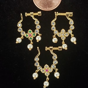 May include: Three gold nose rings with a screw clasp. Each ring is decorated with small clear rhinestones and a dangling pearl. The center ring has a pink and green gemstone flower.