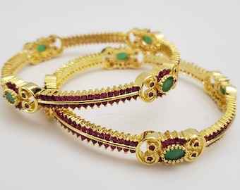 May include: Two gold bangles featuring red and green stones. The bangles are adorned with elaborate designs and have a polished finish.
