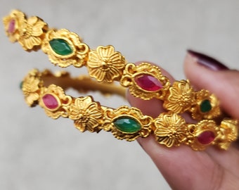 Puede incluir: Un par de brazaletes de oro con intrincados diseños florales y piedras preciosas rojas y verdes.