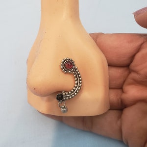 May include: A silver nose ring with a red and black stone. The ring is in the shape of a curved line with a small dangling ball.
