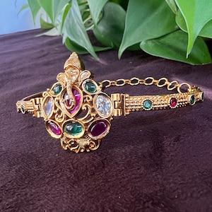 May include: A gold-toned, ornate, adjustable bangle with a detailed elephant design and multi-colored gemstones. The bangle is decorated with green, red, and white stones.