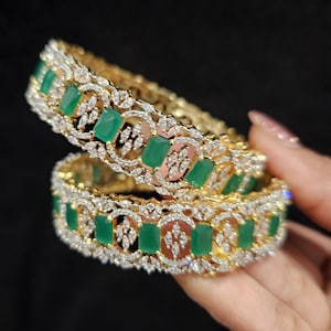 May include: Two gold bangles with a row of emerald green square stones set in a pave diamond design.