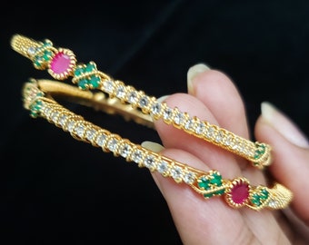 May include: Two gold bangles with white and green stones and a red stone accent.