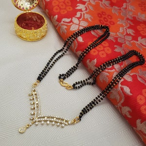 May include: A three-strand black beaded necklace with a gold pendant featuring a V-shaped design with clear rhinestones. The necklace is lying on a white surface with a red and gold patterned fabric in the background.