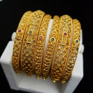 May include: Set of six gold-tone bangles with intricate floral designs and small green and red gemstones.