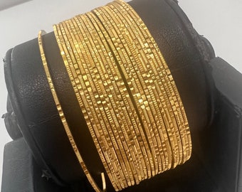 May include: A stack of 18 gold bangles with a textured design. The bangles are arranged in a circular pattern on a black display stand.