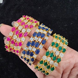 May include: Four gold bangles with white cubic zirconia stones and coloured gemstones. The bangles are stacked on top of each other. The top bangle has pink gemstones, the second bangle has blue gemstones, the third bangle has green gemstones, and the bottom bangle has white gemstones.