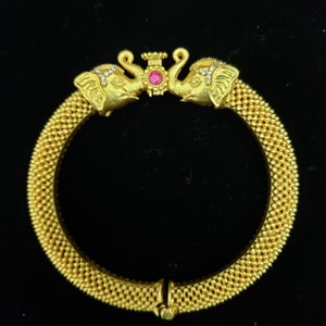 Peut inclure: Un bracelet jonc en or avec deux têtes d'éléphants sur le dessus. Les éléphants se font face et ont une pierre rose au centre. Le bracelet est fait de petites perles rondes.