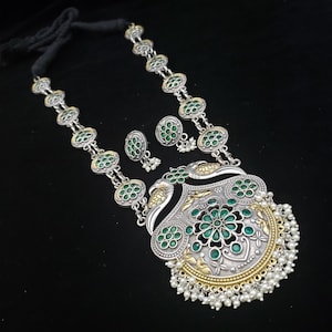 May include: A silver and gold necklace with a large pendant featuring a peacock design and green stones. The pendant is surrounded by a fringe of white pearls. The necklace also includes a pair of matching earrings with green stones.