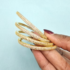 May include: Four gold-colored bangles with a continuous row of small, clear gemstones. The bangles are held against a light blue background. The gemstones sparkle, reflecting light. The bangles are stacked together.
