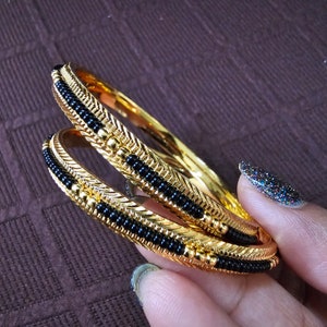 May include: Two gold bangles with black beads. The bangles are stacked on top of each other and have a decorative design.