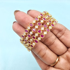 May include: Four gold bangles with alternating ruby-red oval stones and clear, sparkling diamond-like accents. The bangles are held against a light blue background, showcasing their intricate design and vibrant colors.
