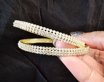 Puede incluir: Dos brazaletes de oro con una fila de piedras brillantes transparentes alrededor de cada brazalete.