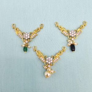 May include: Three gold-coloured jewellery pendants with ornate floral designs. Each pendant features a central cluster of clear crystals and dangling pearls or gemstones. One has a green gemstone, another a black gemstone, and the third has a cluster of pearls.